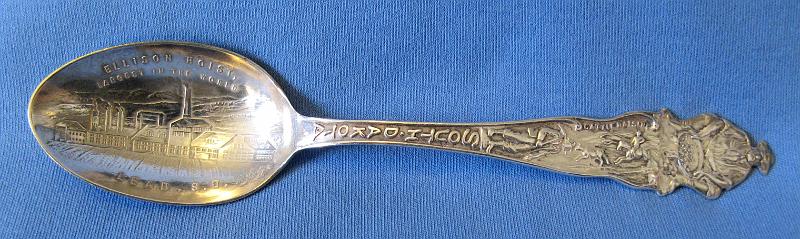 Souvenir Mining Spoon Ellison Hoist Lead SD.JPG - SOUVENIR MINING SPOON ELLISON HOIST LEAD SD - Sterling silver spoon withengraved bowl showing mine buildings and marked ELLISONHOIST LARGEST IN THE WORLD LEAD, S.D. with handle showing miner and gold pan at top, cattle below that, and a miner with pick below that, marked SOUTH DAKOTA mid-handle to bowl, 5 7/8 in. long, reverse marked Sterling  (By 1895, the Homestake Mining Company of Lead, SD recognized the need for a new, larger shaft at the mine due to its growth and expansion and the proximity of the current shafts - the Golden Star, the Golden Prospect, and the B&M - to valuable ore pockets. Construction of the Ellison Shaft on the General Ellison mineral claim began in 1895. Construction on the headframe, hoist house and crusher room got underway in 1897. The shaft was located across from the preexisting shafts on Gold Run gulch.  A 900 foot tramway was built across the gulch to haul ore from the Ellison to the Homestake Mills. The entire shaft, headframe, hoist, crushing plant and tramway were completed on January 1, 1902, costing approximately one million dollars.  The Ellison hoist was the wonder of the age.  It had hoisting engines with a capacity for 3,000 feet; crusher engines with capacity for six No. 6 Gates crushers; a compressor with capacity for 250 drills, and another compressor for the tramway and underground motors. A compressed air motor, hauling 28 steel bottom-dumping cars, containing four tons of ore each, operated between the Ellison hoist and the mills of the Homestake Mining Company, over a steel bridge 100 feet high. The Ellison shaft had three compartments. In two of them double-decked cages were operated, each deck accommodating two cars, holding a ton of ore each; the third compartment contained man-ways, air-pipes, etc.  At the start of the 20th century, the Ellison hoist and a duplicate hoist at the Anaconda copper mine in Butte, MT, along with the great hoist of the Calumet & Hecla copper mine in Michigan, which raised ore from a shaft more than one mile in depth, were the most powerful mine hoists known. The Ellison hoist had no peer on a gold mine anywhere in the world. The hoist building, housing the machinery of the Ellison equipment, was 350 feet long by 100 feet in width, and 80 feet in height over the central portion. The hoist engine was built by the Union Iron Works of San Francisco, CA.  It had two steam cylinders 30 by 72 inches and two steel reels 16 feet in diameter, which wound and payed out a flat steel cable seven inches wide by a full inch in thickness. The two cables were 3000 feet in length each.  The hoist engine was partitioned off in the big building with a glass front toward the shaft. In this compartment were placed two small air compressor engines and auxiliaries for handling and governing the movements of the great machine. The clutches, post brakes, disc brakes and reverse gear were operated by compressed air and were entirely controlled by a set of three levers and one pedal, conveniently placed on an elevated platform from which the engineer had a full view of all the machinery and the shaft. When ore was supplied sufficient to keep the hoist moving, eight tons of ore were raised to the surface every two minutes; three top men dumped the cars, feeding the ore to four No. 6 Gates crushers and returned the empties to the cage. In operation, 5,760 tons of ore were moved every 24 hours. The boiler capacity furnishing the steam power for the hoist consisted of eight Scotch marine boilers with a total capacity of 1,600 horse power. These boilers were made in the shops of W. J. Solbcrg & Son, of La Crosse, WI.  Over the next several decades, many buildings were constructed around the headframe to support operations.  On the evening of July 10, 1930, tragedy struck the Ellison Shaft.  A fire ignited from overheated pipes in the air compressor room shortly before 8 pm. Within minutes, the headframe and many smaller buildings nearby were rapidly burning.  Two men trapped in the shaft were killed. The loss of these two lives and the destruction of the Ellison Shaft were truly devastating.  The hoist, headframe and shaft were a mass of wreckage, debris, and steel beams.  The rebuild started almost immediately and was complete in February 1931.)